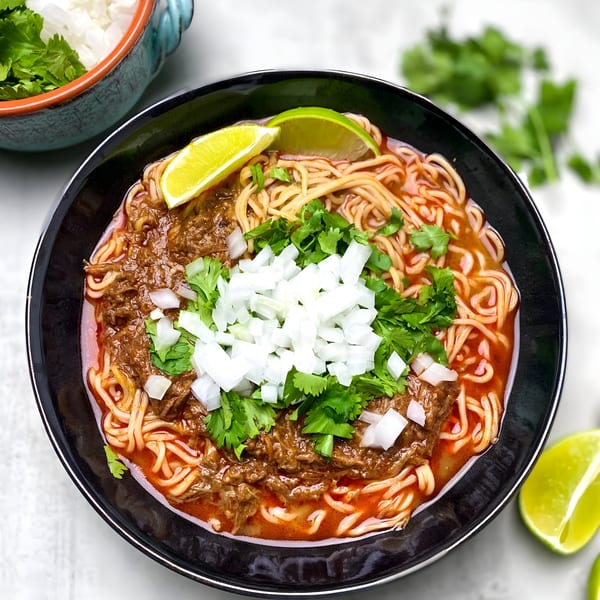 Chido's Tacos birria ramen with shredded beef, noodles, cilantro, onions, and lime wedges.