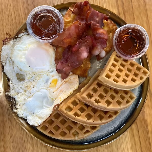 A breakfast plate with fried eggs, bacon, hash browns, waffles, and two cups of syrup.