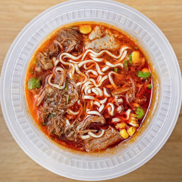 Overhead view of a birria ramen bowl with noodles, shredded beef, corn, and cilantro in a rich red broth.