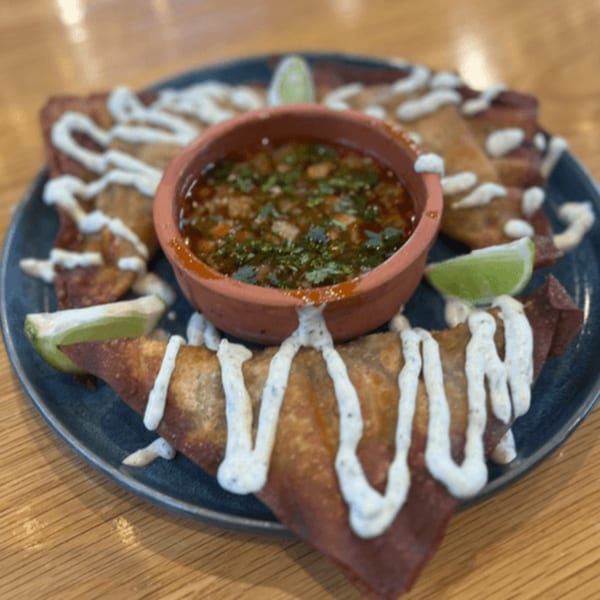 Crispy Chido's Tacos with white sauce drizzle, lime wedges, and a bowl of salsa on a dark plate.