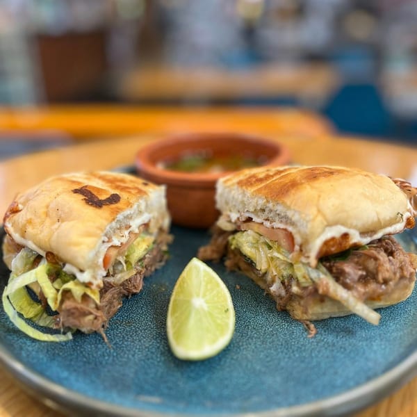 A Mexican torta sandwich, cut in half, with a lime wedge and a small bowl of soup.